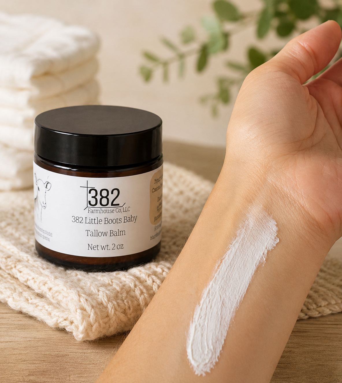 A jar of 382 Farmhouse Grove 382 Little Breeze Baby Tallow Balm sits on a knitted beige cloth next to a person's forearm showing a thick white balm swatch.